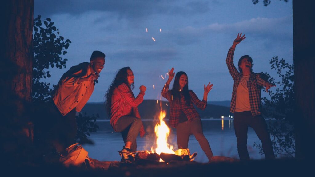 A group of friends dancing and singing around a campfire by the lake at night.